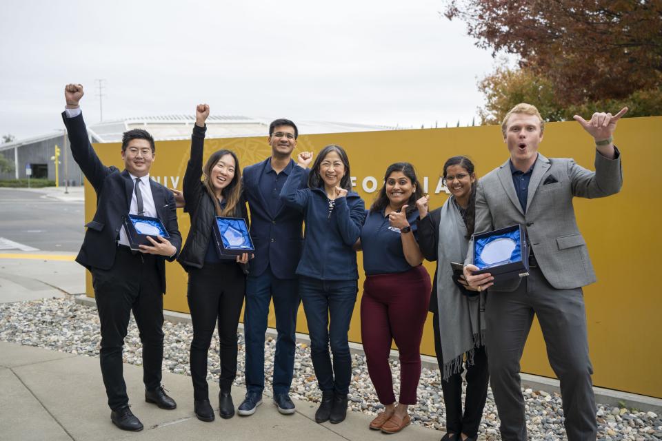 Group of individuals standing in front of the UC Davis sign with their hands raised celebrating